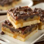 Stacked Millionaire’s Shortbread Bars with a buttery shortbread base, gooey caramel, and a rich chocolate topping, garnished with chocolate chips.